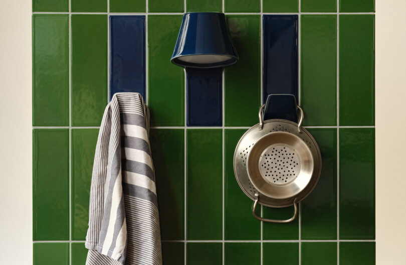 A blue cup, a striped towel, and a metal colander hang on hooks attached to a green tiled wall.