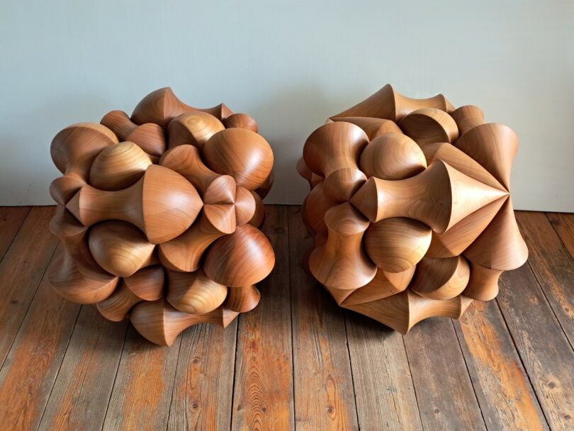 Two wooden geometric sculptures with protruding rounded and conical shapes sit on a wooden floor against a plain white wall.