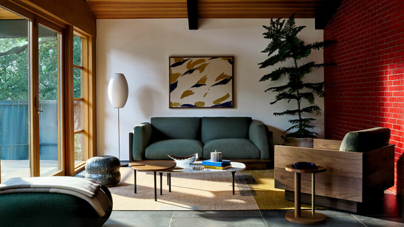 Modern living room with dark green sofa, wooden armchair, abstract wall art, potted plant, and large windows. A striking red brick accent wall.
