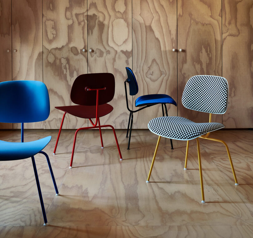 Four modern chairs in different colors and patterns are arranged on a plywood floor in front of a wood-paneled wall with cabinets.