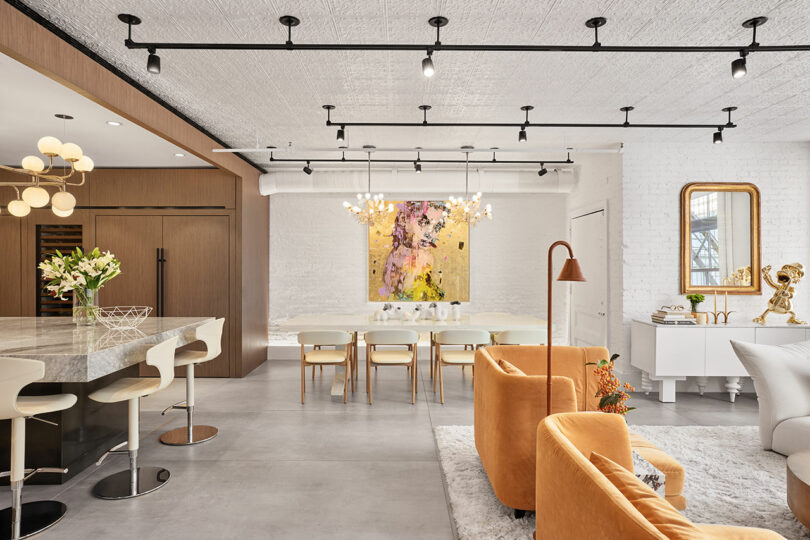 Modern open-plan living space with a kitchen, dining area, and lounge featuring neutral tones, gold accents, contemporary furniture, and colorful artwork on a white brick wall