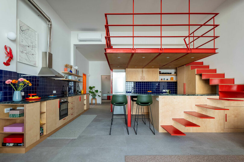 Modern kitchen with light wood cabinets, blue tile backsplash, central island with green stools, and red metal stairs leading to a loft space