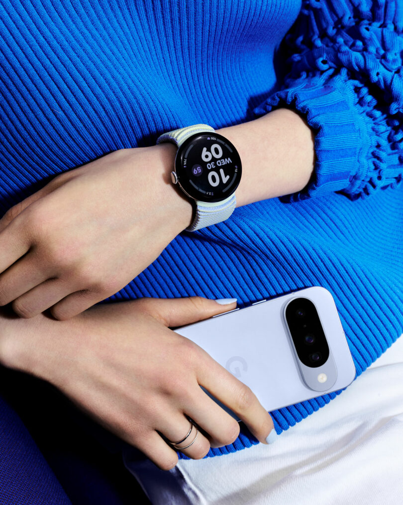 A person wearing a blue long-sleeve top rests their hand, with a smartwatch displaying time and date, on a white smartphone with dual cameras—showcasing the seamless integration of modern technology.