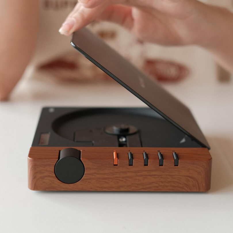 A person’s hand is about to close the lid of a modern, wood-finish CD player, showcasing sleek technology with visible control buttons and a round knob.