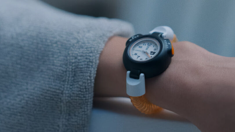 A person wearing a watch with a white face, black bezel, and yellow wristband—a sleek piece of technology—with the sleeve of a light gray textured garment partially visible.
