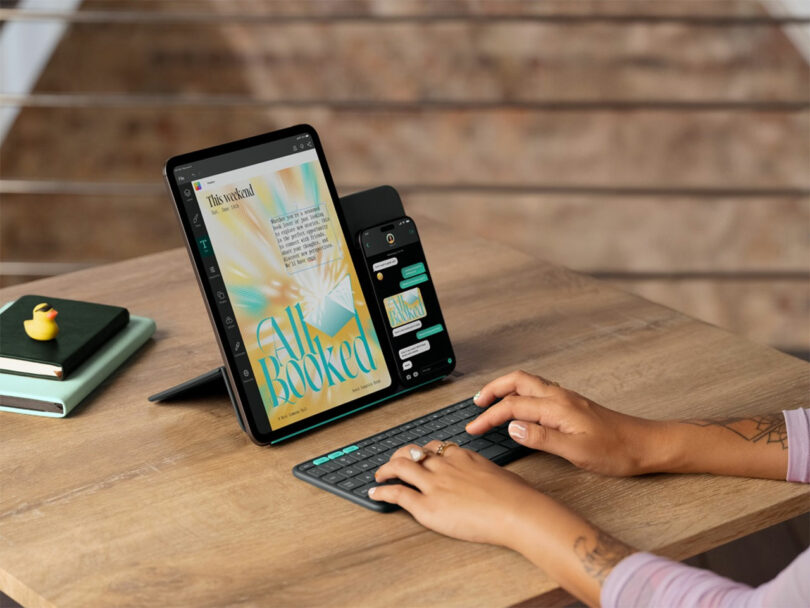 A person types on a wireless keyboard connected to tablet and smartphone, showcasing seamless technology, both propped up on a desk with notebooks and a duck-shaped eraser nearby.