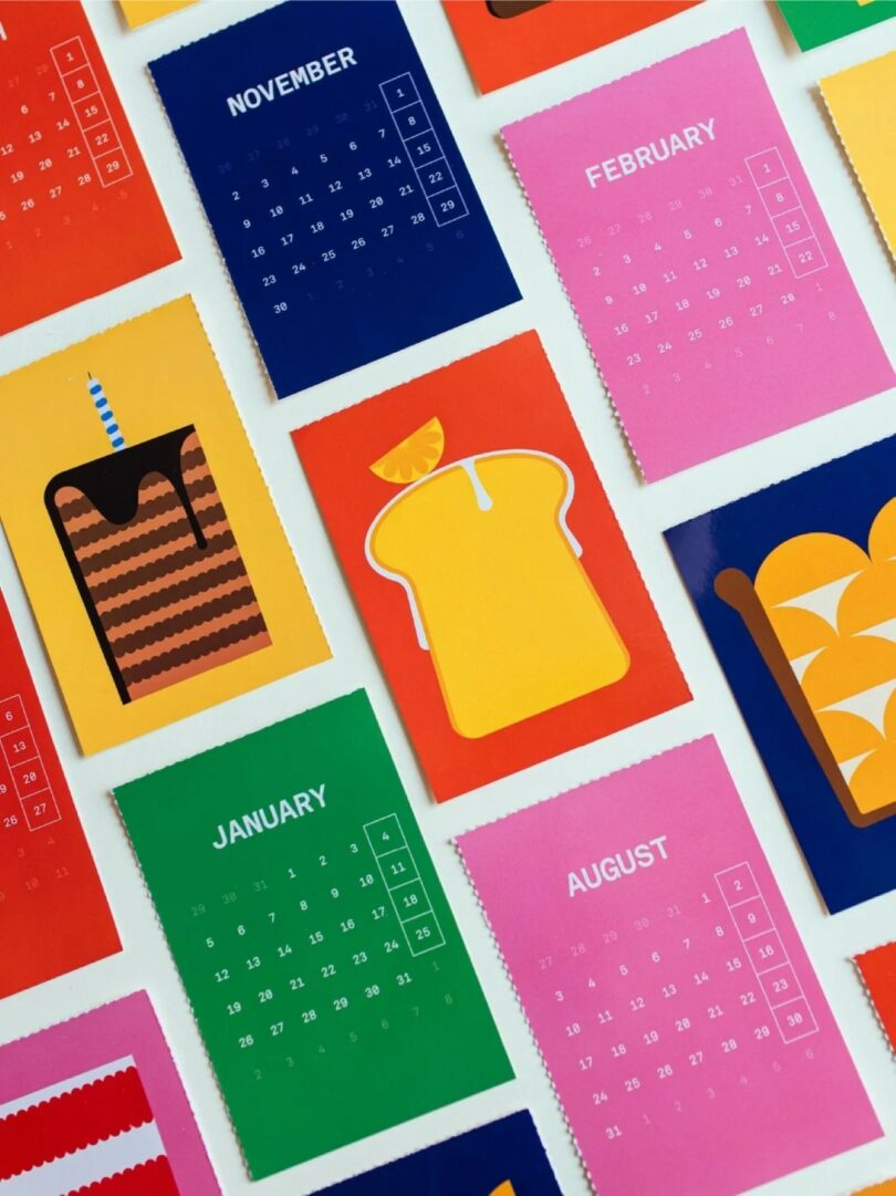 Colorful 2026 calendars, with pages for each month arranged diagonally on a white surface, feature simple food illustrations.