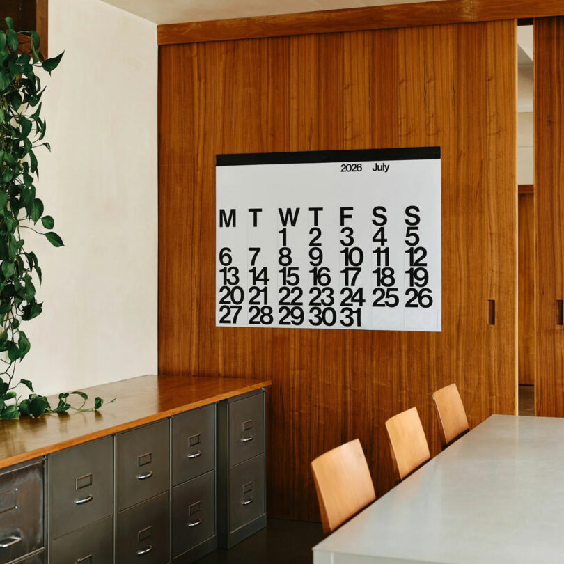 A large 2026 calendar displaying July hangs on a wooden wall above metal filing cabinets and a conference table with wooden chairs; a leafy plant sits to the left.