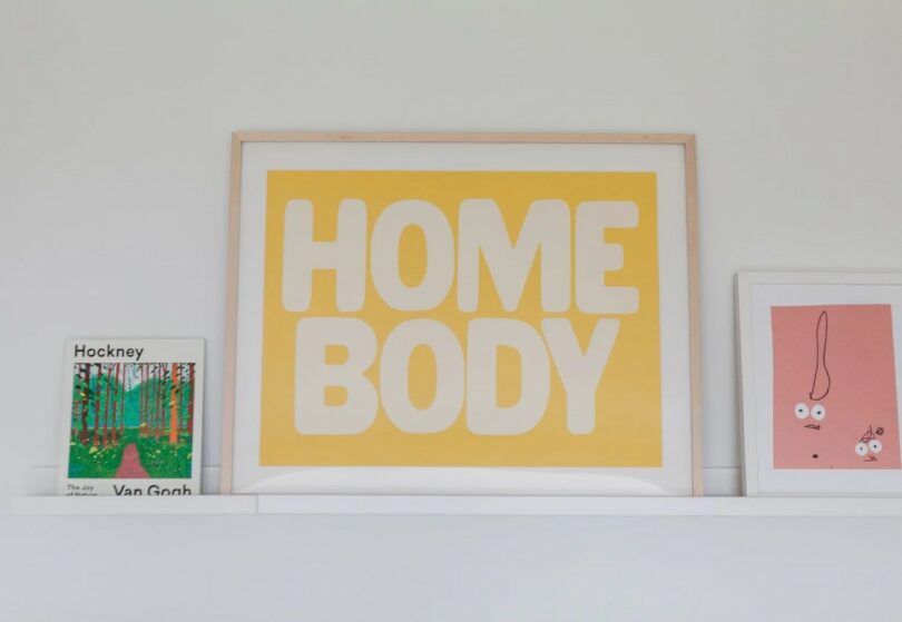 A framed print with the words "HOME BODY" in large yellow and white letters sits on a shelf between a Van Gogh book and a pink art print with abstract face drawings