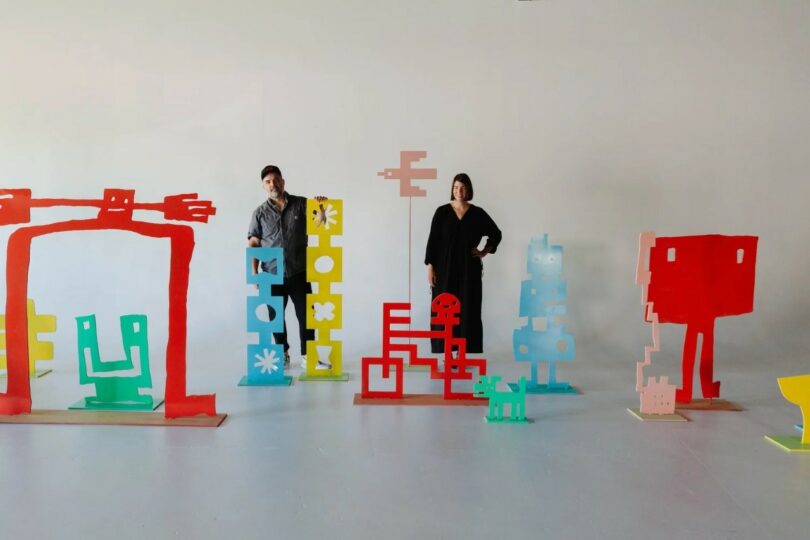 Two people stand in a studio among colorful abstract sculptures made of flat, painted shapes in red, blue, yellow, and green