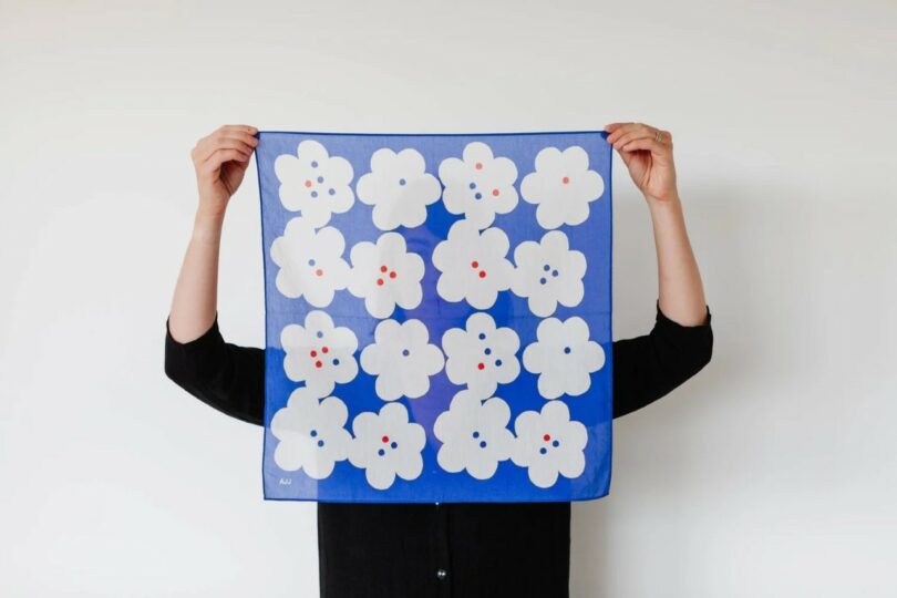 Person in a black shirt holds up a blue square cloth with a pattern of white flowers, each with colorful dots at the center, against a plain white background