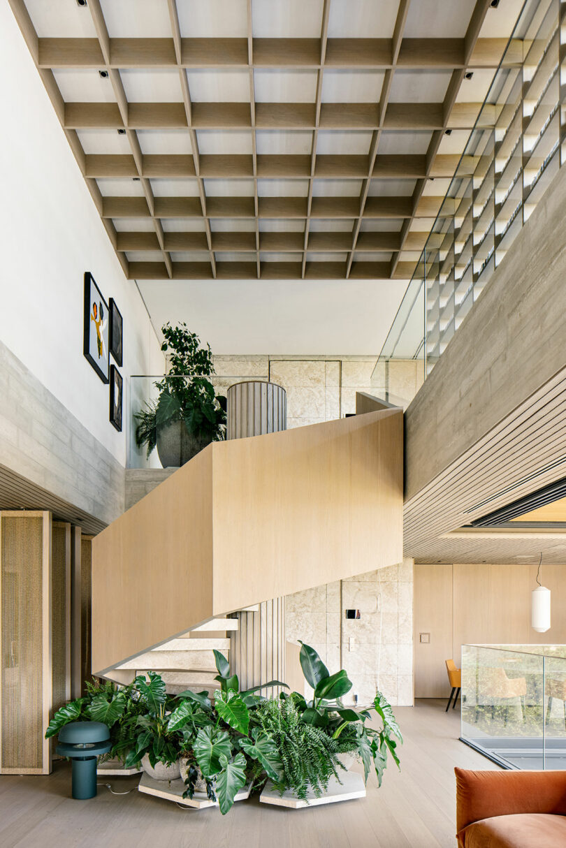 Modern interior with a central wooden staircase, large potted plants, and natural light. Features a grid ceiling, glass railing, and minimalist decor. Artworks visible on the upper wall.