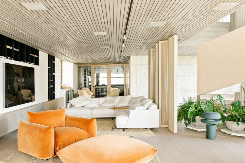Modern living room with a large white sectional sofa, orange velvet chair and ottoman, indoor plants, built-in shelving, and wood-paneled ceiling and floors.