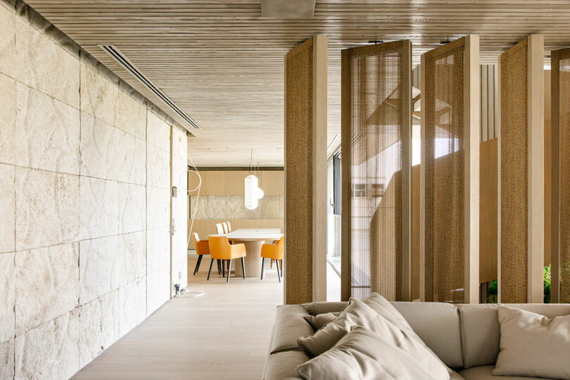 Modern living space with beige sofa, textured stone wall, wooden partition panels, and a dining area with orange chairs and a light wood table in the background.