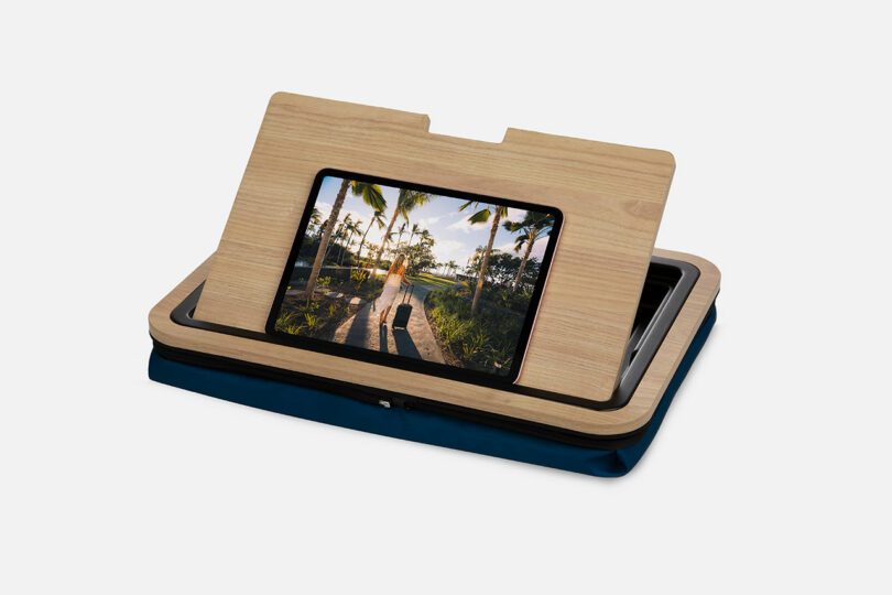 A wooden tablet stand holds a tablet displaying a beach scene, all set atop a navy blue zippered pouch against a plain white background.