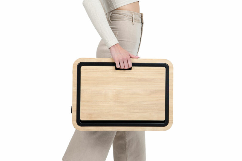A person holding a rectangular wooden cutting board with a black border and a handle, standing against a white background.