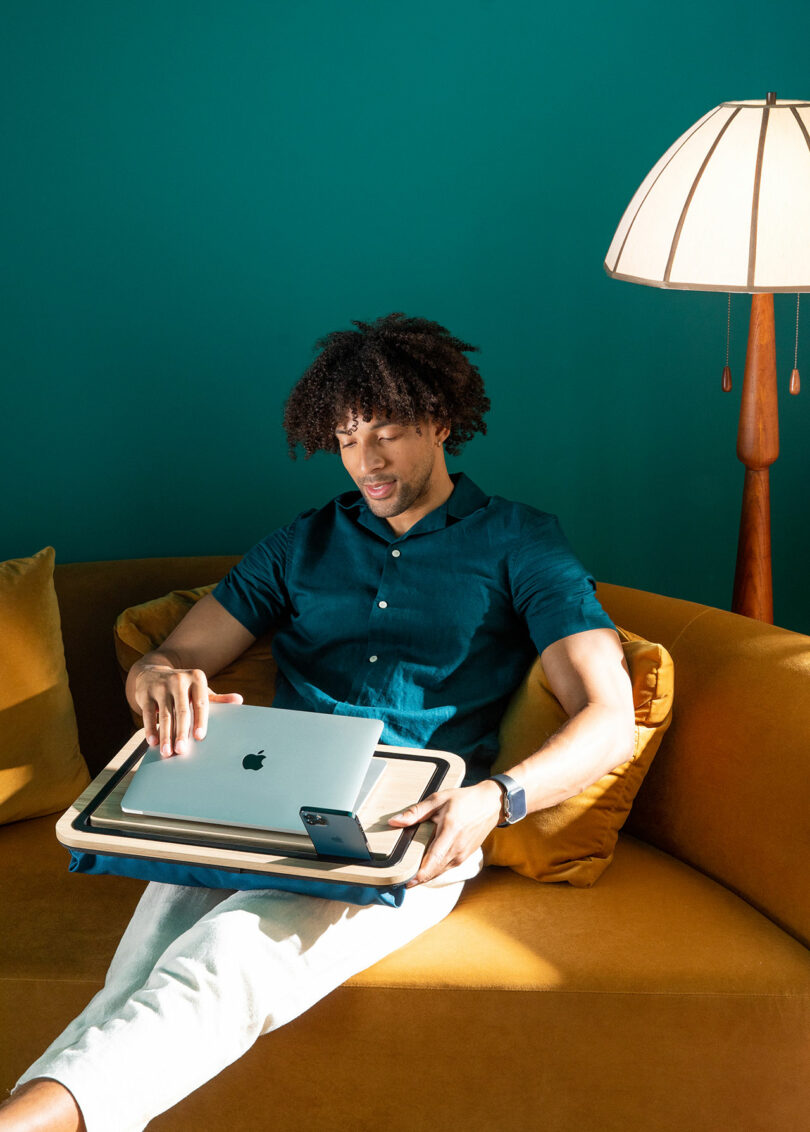 A man sitting on a mustard yellow couch holds a MacBook, iPad, and iPhone on his lap, with a lamp and teal wall in the background.