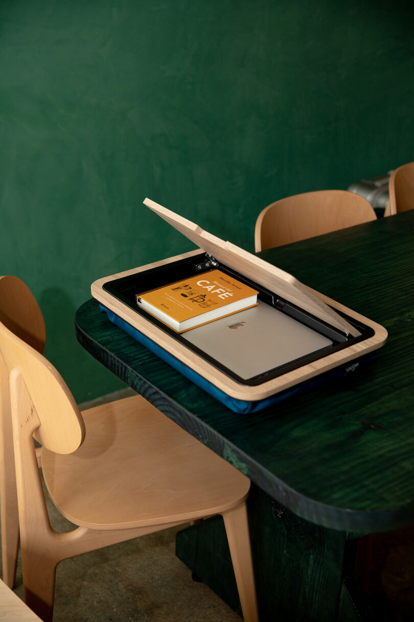 A wooden box with a partially open lid contains a closed silver laptop and a yellow book, placed on a dark green table next to light wooden chairs.