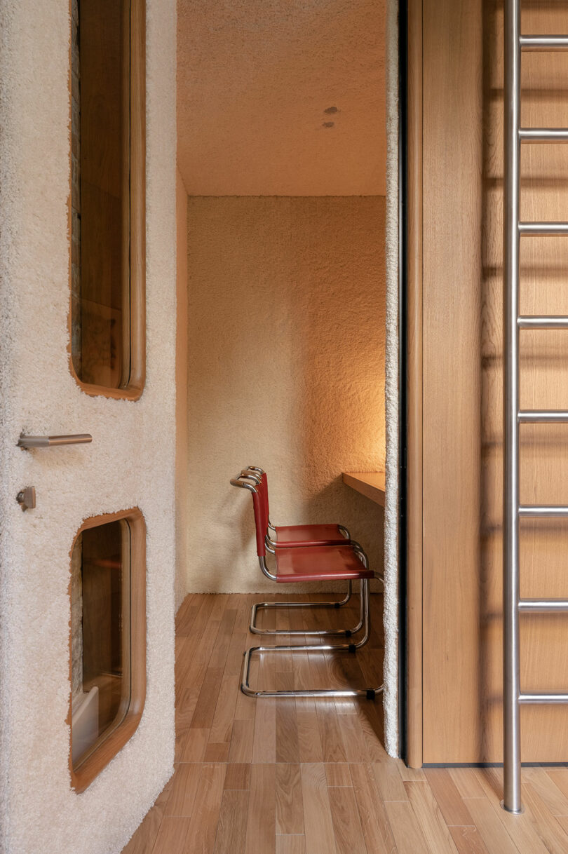A red and chrome chair sits at a wooden desk in a small, textured-walled room with wooden flooring and a metal ladder beside a door.