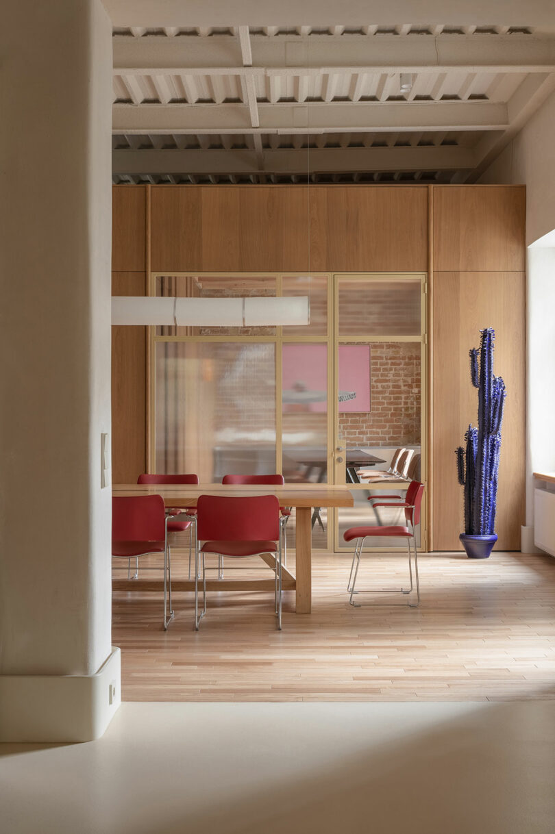 A modern office space with a wooden table, red chairs, a glass partition, a blue potted cactus, and natural light from a window.