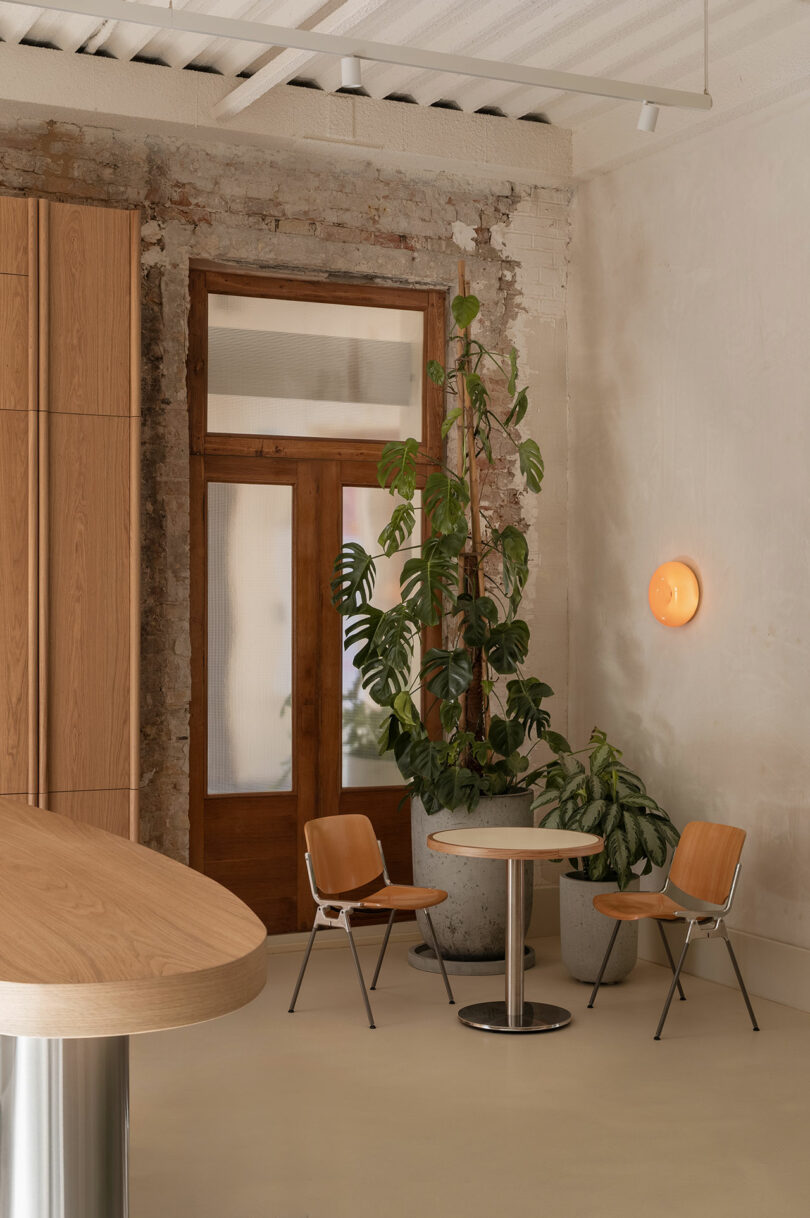 A small round table with two wooden chairs sits near a large potted plant and a wooden-framed glass door in a minimalist room with exposed brick and a wall-mounted light.