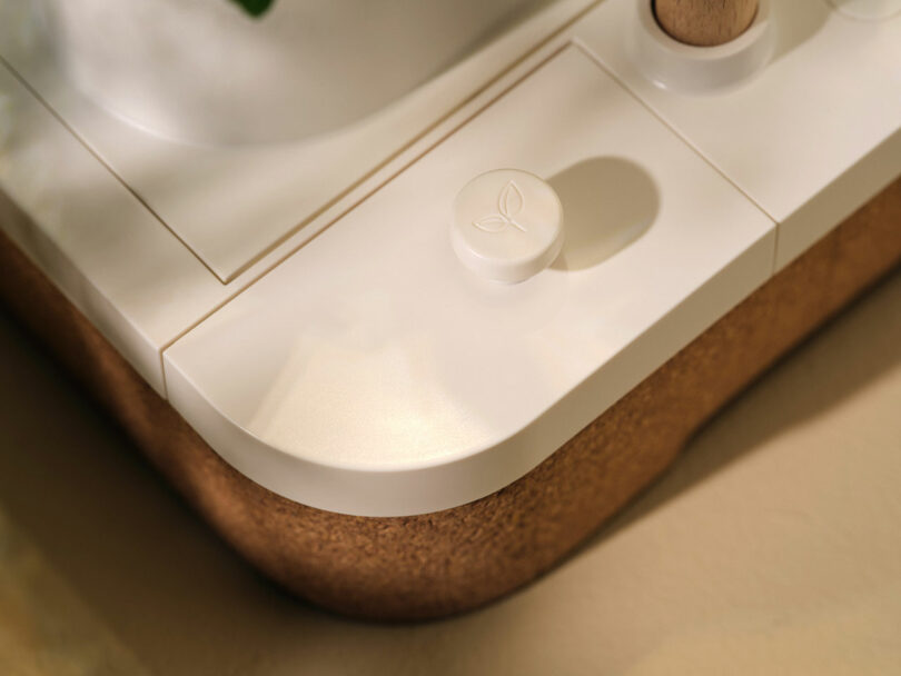 A close-up of a modern white and cork device with a round button featuring a leaf symbol, placed on a light-colored surface.