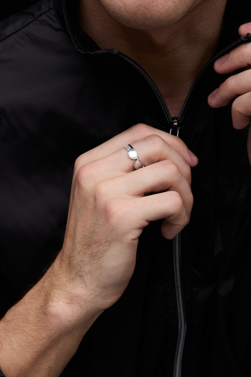 A person wearing a black zip-up jacket holds the zipper with one hand, displaying a silver ring on their finger.
