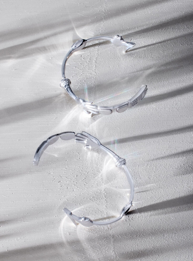 Two silver bracelets with hand-shaped ends placed on a textured white surface, with light creating soft shadows and reflections around them.