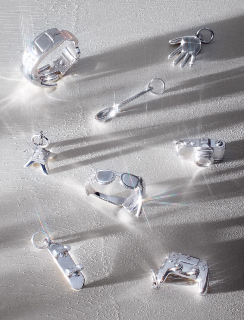 A collection of shiny silver charms arranged on a textured surface, including a bracelet, glove, spoon, star, sunglasses, camera, skateboard, and game controller.