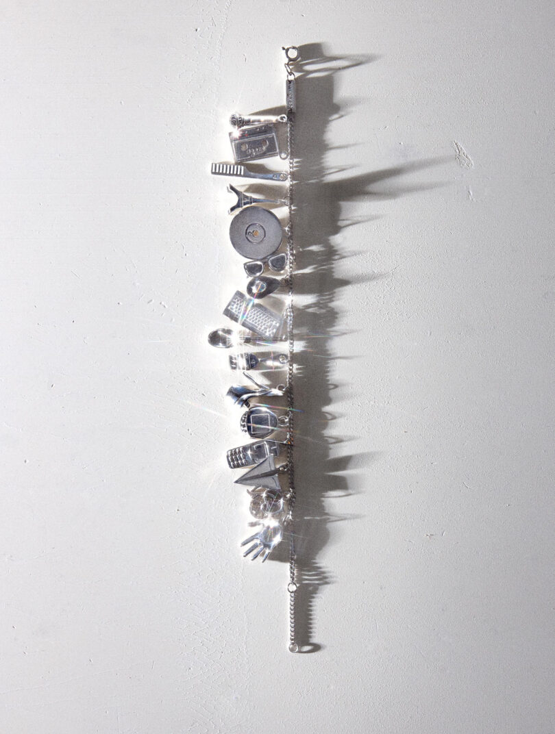 A silver charm bracelet with various detailed charms, including a camera, heart, book, and key, is laid flat on a light gray surface with shadows visible.