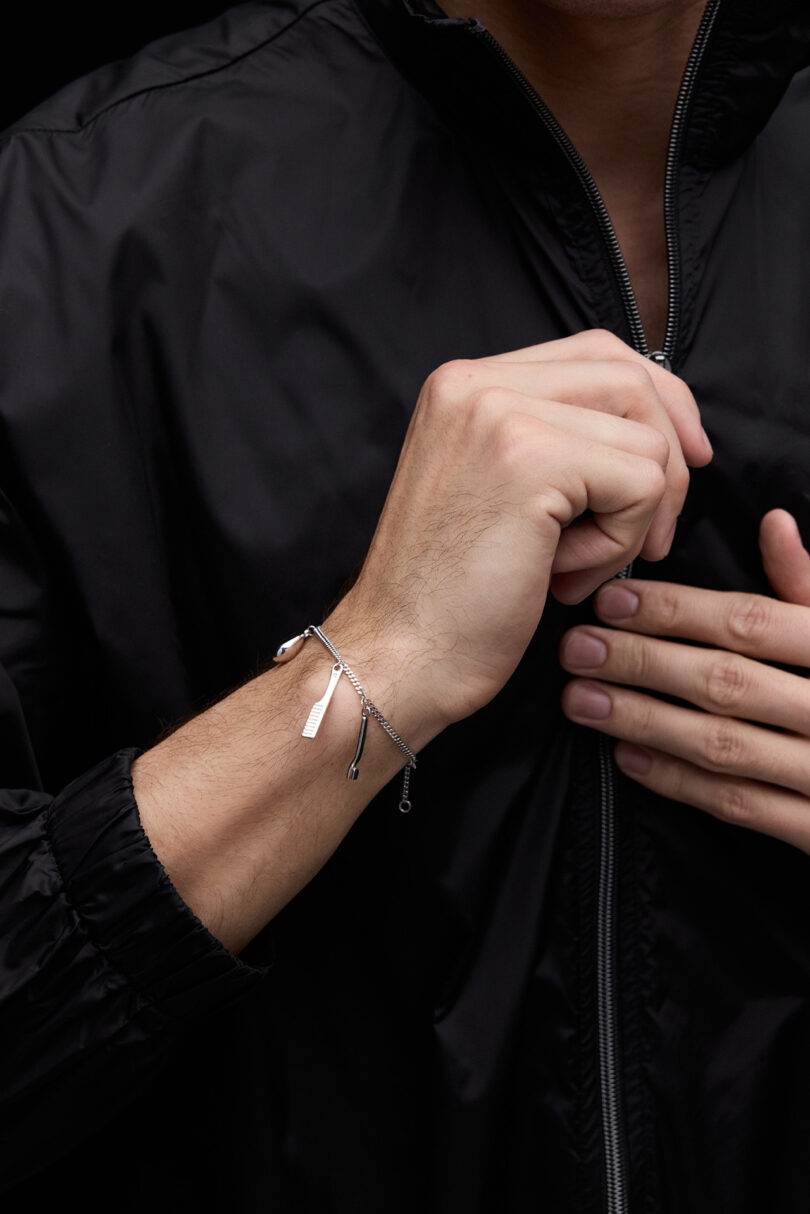 A person wearing a black zip-up jacket displays a silver chain bracelet on their wrist, with one hand held to their chest.