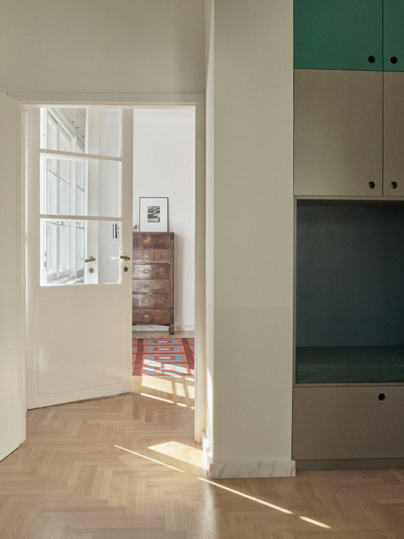 Sunlight enters a minimalist room with parquet flooring, a partially open door, built-in cabinets, and a chest of drawers visible in the adjacent room.
