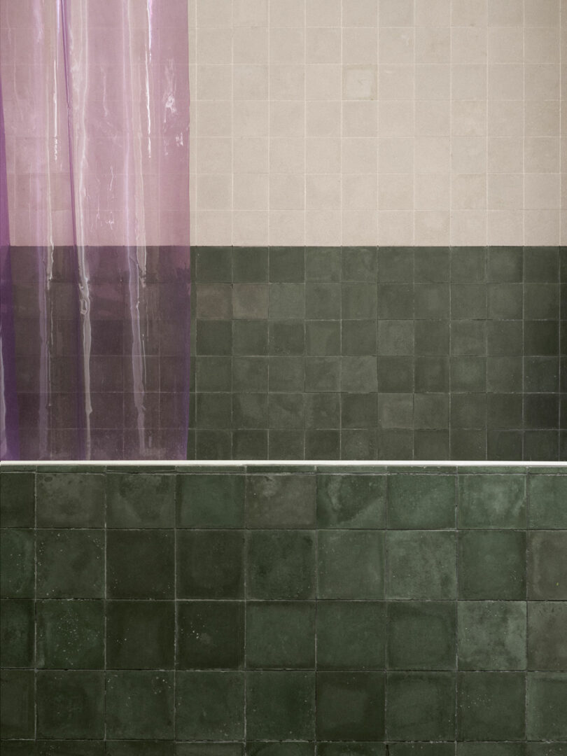A pink shower curtain partially covers a wall with light beige and dark green square tiles above a dark green tiled bathtub.