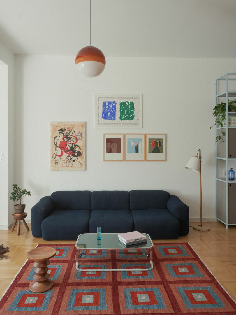 A modern living room with a dark blue sofa, colorful geometric rug, glass coffee table with books, framed art on the wall, a floor lamp, and potted plants.