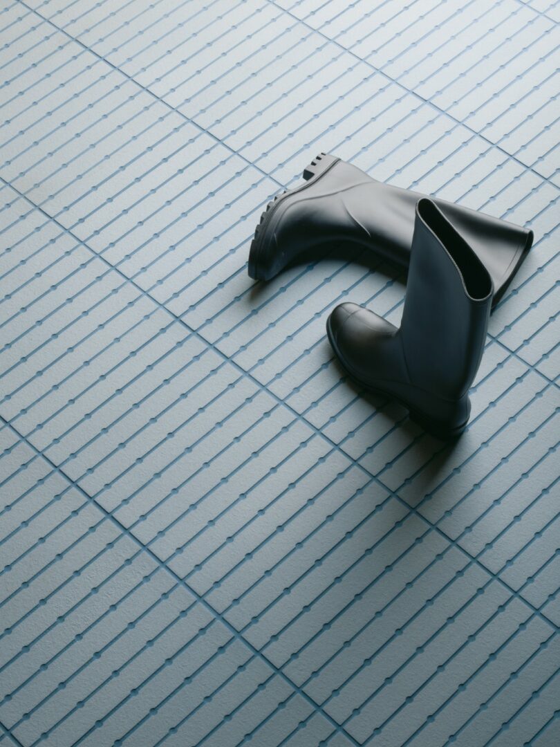 A pair of black rubber boots sits on a light blue tiled floor with a geometric grid and dot pattern