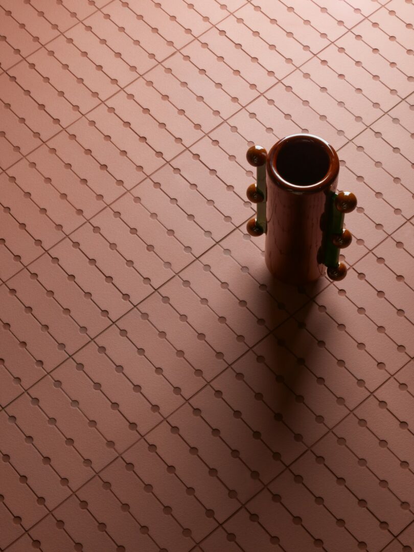 A cylindrical vase with rounded handles stands on a pink patterned surface, casting a shadow to the right