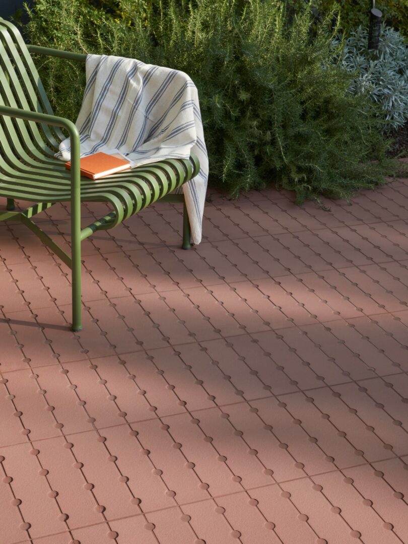 A green metal chair with a striped cloth and orange notebook on its armrest is placed on a patio with interlocking red tiles, next to green plants