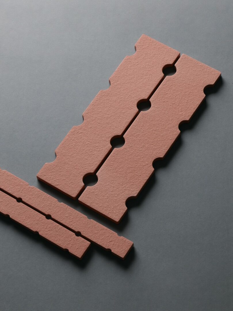Three textured, reddish-brown rectangular tiles with interlocking notches and circular cutouts are arranged on a gray surface