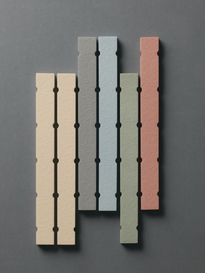 Five vertical, textured rectangular tiles in beige, pale yellow, light blue, green, and red are arranged side by side on a dark grey background