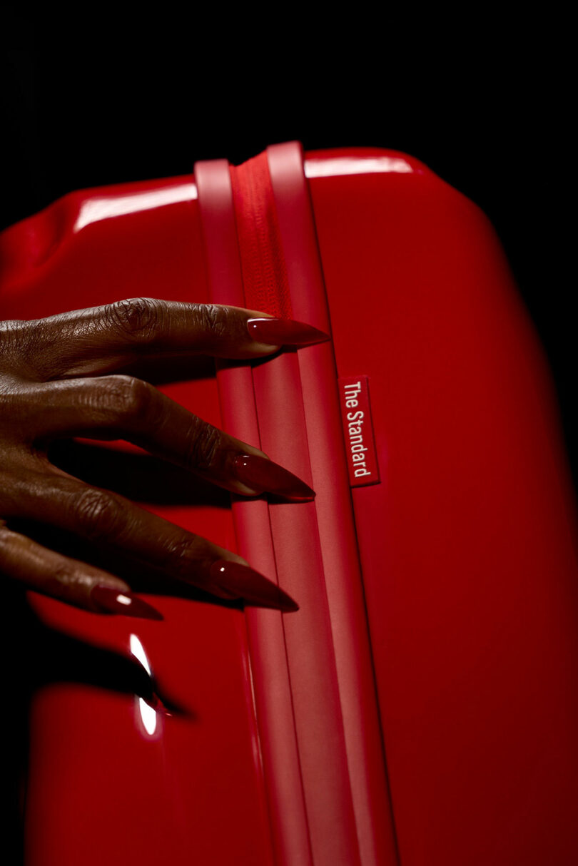 A hand with long, red nails rests on a glossy Crash Baggage suitcase with a small label that reads "The Standard" against a dark background.