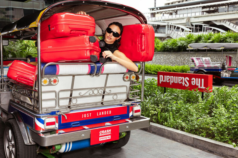 A woman wearing sunglasses leans out of the back of a tuk-tuk loaded with red Crash Baggage suitcases, parked near a hotel with a sign reading "The Standard.