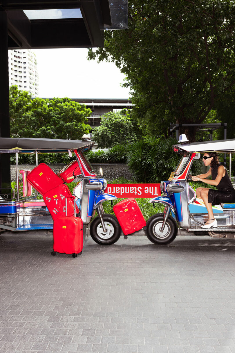 A woman in sunglasses sits in a tuk-tuk with several red Crash Baggage suitcases stacked between two tuk-tuks outside The Standard hotel.