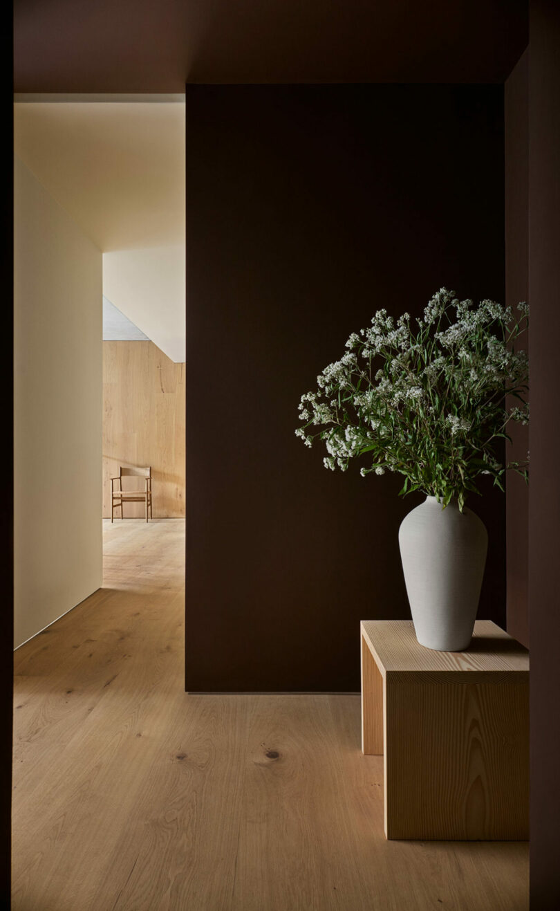 A white vase with white flowers sits on a wooden bench in a minimalist hallway with dark brown and light wood walls and flooring. A wooden chair is visible in the next room.