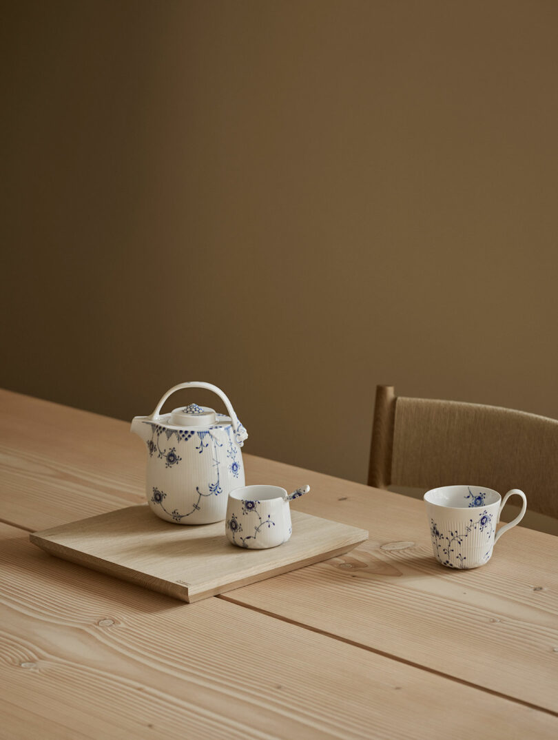 A white ceramic teapot, sugar bowl, and teacup with blue floral patterns are arranged on a wooden table with a beige background.