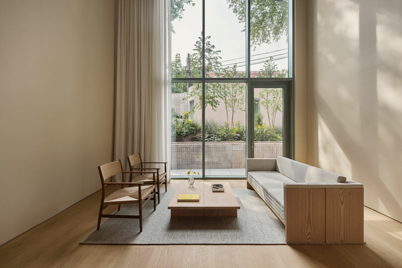 Minimalist living room with light wood furniture, a gray rug, and large floor-to-ceiling window overlooking a garden with trees and shrubs.