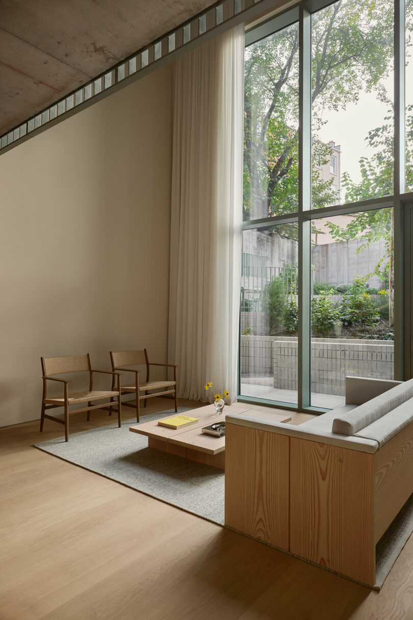 Minimalist living room with wood furniture, floor-to-ceiling windows, neutral tones, and a view of a garden outside. Natural light fills the space.