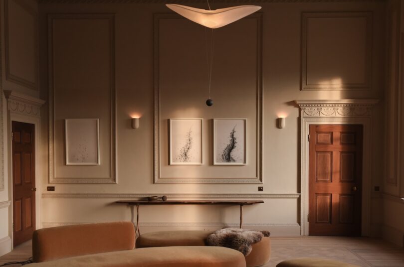 A minimalist living room with beige walls, three abstract framed artworks, a wooden door, a curved sofa, and a modern pendant light.