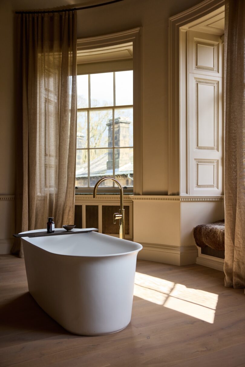 A modern white bathtub with a gold faucet stands by large windows with beige curtains in a sunlit, elegant bathroom.
