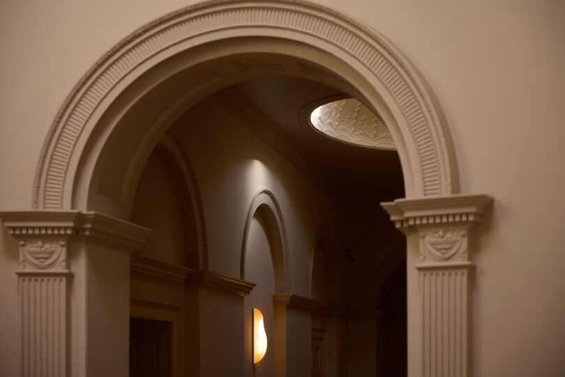 An ornate, cream-colored interior archway with decorative molding and columns, illuminated by wall sconces and a small round skylight above.