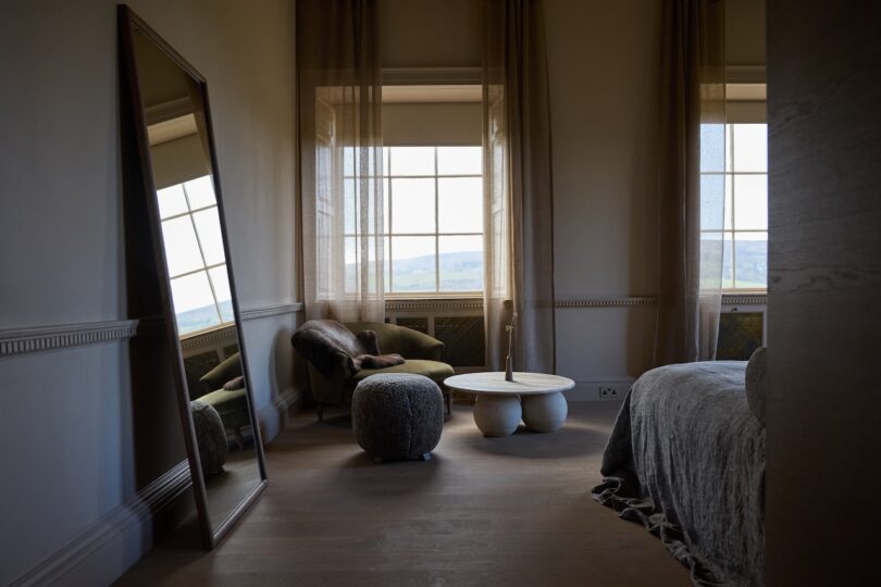 A softly lit bedroom with a large mirror, armchair, round ottoman, two round coffee tables, and sheer curtains over a window.